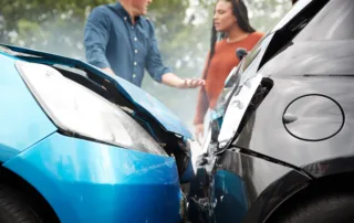 Two cars, blue and black, with damaged hoods after a head-on collision. A man and woman in the background discuss the accident, appearing concerned.