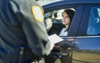 A woman sitting in her car looking up at a police officer holding a notebook during a traffic stop for driving uninsured.