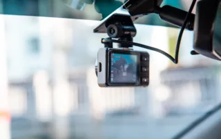 Compact dashcam mounted on a car windshield showing live street view with side buttons and a visible power cable.